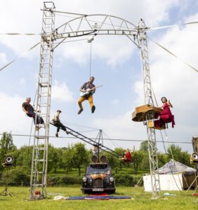 Acrobats perform suspended on a metal frame above a vintage car, with a blue sky and grassy field in the background.